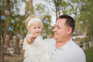 Vater, der ein kleines Mädchen hält, das Blasen draußen im Park genießt foto