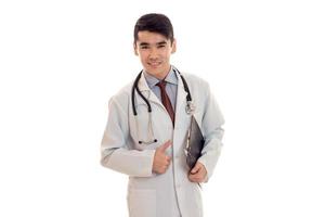 junger eleganter Arzt in Uniform mit Stethoskop lächelnd vor der Kamera mit Brett in den Händen isoliert auf weißem Hintergrund foto