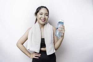 sportliche asiatin posiert mit einem handtuch auf der schulter und hält eine flasche wasser, lächelt und entspannt sich nach dem training foto