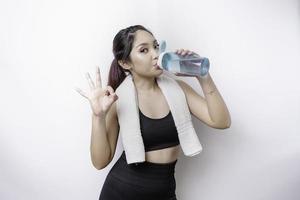 sportliche asiatin posiert mit einem handtuch auf der schulter und trinkt aus einer flasche wasser, lächelt und entspannt sich nach dem training foto