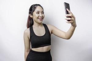 ein Porträt einer sportlichen asiatischen Frau, die eine Sportkleidung trägt und ihr Telefon hält, isoliert durch weißen Hintergrund foto