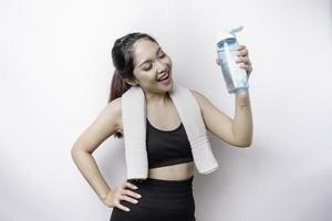sportliche asiatin posiert mit einem handtuch auf der schulter und hält eine flasche wasser, lächelt und entspannt sich nach dem training foto
