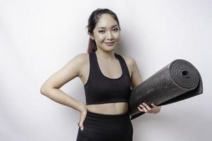 lächelnde sportliche asiatische frau, die sportkleidung trägt, hält yogamatte auf lokalisiertem weißem hintergrund. foto