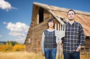 Biracial chinesisches und kaukasisches junges Paar, das eine Holzgabel vor einer rustikalen Scheune auf dem Land hält. foto