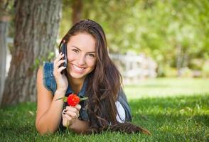 Attraktive, glückliche, gemischte Abstammung, junge Frau, die am Handy außerhalb der Verlegung im Gras spricht foto