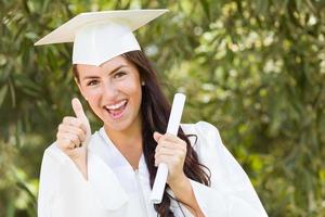 Mixed Race Daumen hoch Mädchen feiert Abschluss draußen in Mütze und Kleid mit Diplom in der Hand foto