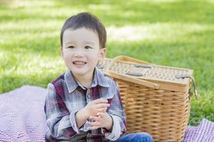 junger Mischlingsjunge, der im Park in der Nähe von Picknickkorb sitzt foto