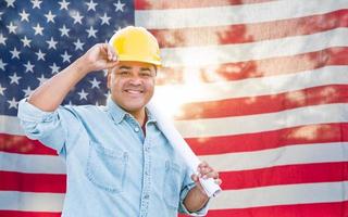 Hispanischer männlicher Auftragnehmer mit Blaupausenplänen, die Schutzhelm vor amerikanischer Flagge tragen foto