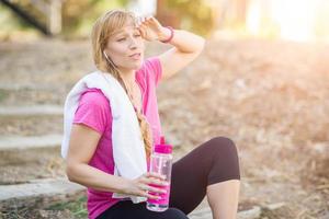 junge fitte erwachsene frau im freien mit handtuch und wasserflasche in trainingskleidung, die musik mit kopfhörern hört. foto