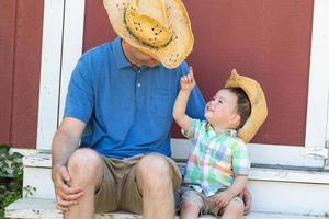 verspielter junger kaukasischer vater und gemischtrassiger chinesischer sohn mit cowboyhüten foto