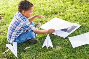 afroamerikanischer und mexikanischer junge, der lernt, wie man papierflugzeuge draußen auf dem gras faltet foto
