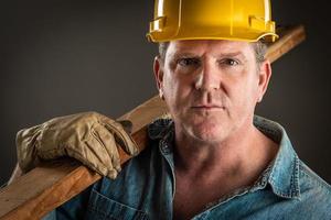 seriöser Bauunternehmer mit Schutzhelm, der ein Holzbrett mit dramatischer Beleuchtung hält. foto