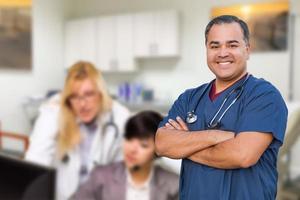 gutaussehender hispanischer arzt oder krankenschwester, der in seinem büro steht, hinter dem mitarbeiter arbeiten foto