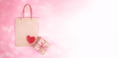 valentinstag festliches banner. diy verpackte geschenkbox, papiertüte, rotes weidenherz auf rosa bokeh hintergrund mit herzen. foto