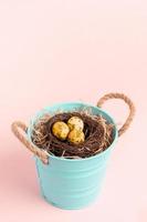 kleiner türkisfarbener Eimer mit Nest aus farbigen goldenen Wachteleiern auf Pastellrosa. Attrappe, Lehrmodell, Simulation. foto
