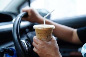 asiatische fahrerin hält eiskaffeetasse zum trinken im auto, gefährlich und riskiert einen unfall. foto