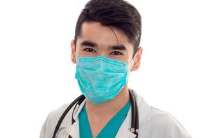 Porträt eines jungen brünetten Arztes in weißer Uniform und Maske mit Stethoskop, der isoliert im Studio in die Kamera blickt foto