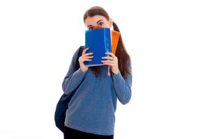 Junge, lustige, brünette Studentin mit Rucksack auf den Schultern, die ihr Gesicht hinter Büchern versteckt, die auf weißem Hintergrund isoliert sind foto