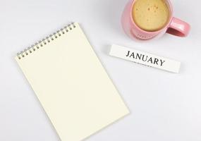 flache lage aus leerem papiernotizbuch, holzkalender januar und rosa tasse kaffee auf weißem hintergrund. foto