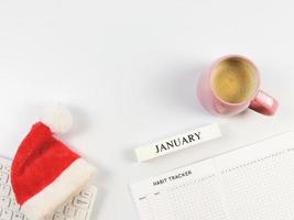flache lage des gewohnheitstrackerbuchs, holzkalender januar, rote weihnachtsweihnachtsmütze auf weißer computertastatur und rosa tasse kaffee auf weißem hintergrund mit kopienraum. foto