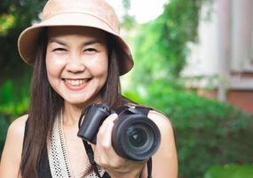 asiatische frau, mit hut und schwarzem oberteil ohne ärmel, steht im garten, hält dslr-kamera und lächelt glücklich. foto