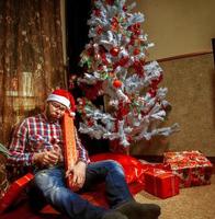 Betrunkener Nerd mit Weihnachtsmütze, der mit vielen Geschenken unter dem Weihnachtsbaum liegt foto