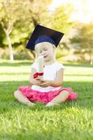 kleines Mädchen im Gras, das eine Abschlusskappe trägt, die ein Diplom mit Band hält foto