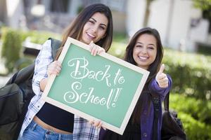 Studentinnen gemischter Rassen, die eine Tafel mit dem Rücken zur Schule halten foto