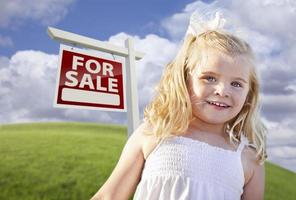 Lächelndes süßes Mädchen im Feld mit Immobilienschild zum Verkauf foto