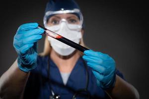 Labormitarbeiterin mit medizinischer Gesichtsmaske hält Reagenzglas mit Blut vor dunklem Hintergrund foto
