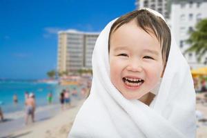 Fröhlicher süßer gemischter chinesischer und kaukasischer Junge am Strand von Waikiki, Hawaii, in ein Handtuch gewickelt foto