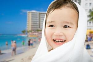 Fröhlicher süßer gemischter chinesischer und kaukasischer Junge am Strand von Waikiki, Hawaii, in ein Handtuch gewickelt foto