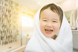 Fröhlicher süßer gemischter chinesischer und kaukasischer Junge im Badezimmer, eingewickelt in ein Handtuch foto