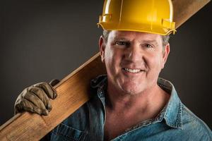 lächelnder Bauunternehmer mit Schutzhelm, der ein Holzbrett mit dramatischer Beleuchtung hält. foto