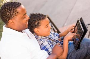 Afroamerikanischer Vater und Mischlingssohn mit Computertablett auf Bank im Park foto