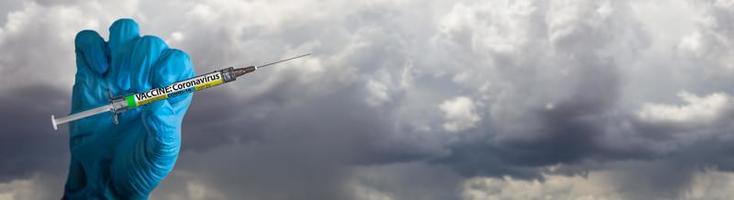 arzt oder krankenschwester, die einen chirurgischen handschuh tragen, der eine medizinische spritze mit einem coronavirus-covid-19-impfetikettenbanner aus wolken hält foto