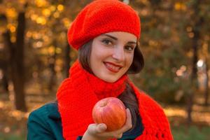 Schönes junges Mädchen in einem roten Schal und Hut lächelnd mit einem Apfel in der Hand, Nahaufnahme foto