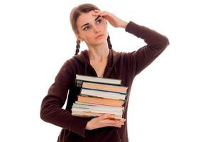 Müde Studentin in brauner Sportkleidung mit vielen Büchern in der einen Hand und der zweiten auf dem Kopf, die isoliert auf weißem Hintergrund posiert foto