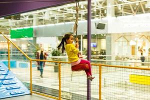 entzückendes kleines Mädchen, das glücklich lacht und kopfüber auf einer Bungee-Schaukel auf dem Spielplatz in Gelb reitet. Glück, Freiheit, Genuss, Gesundheit. foto