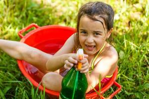 kindliche Entwicklung im Einklang mit der Natur. Kleines schönes Mädchen, das Spaß mit Wasser spielt und im Freien auf dem Gras badet, in Vintage-Waschschüssel-Hygiene, glückliche Kindheit, Naturkonzept foto