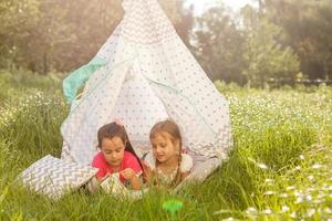 Zwei glückliche lachende kleine Mädchen im Campingzelt auf dem Löwenzahnfeld foto