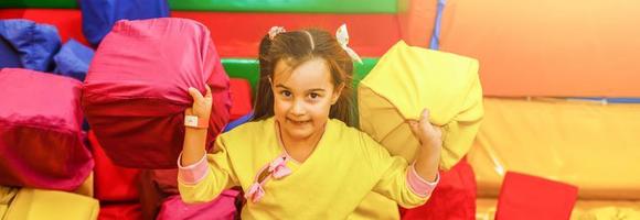 Fröhliches kleines Mädchen, das zum Geburtstag mit weichen Würfeln im trockenen Pool des Spielkinderzimmers spielt. Unterhaltungszentrum. Indoor-Spielplatz in Schaumgummigrube im Trampolin foto