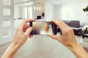 zwei hände, die ein mobiles smartphone halten und ein foto in einem modernen luxuswohnzimmer und einer küche machen