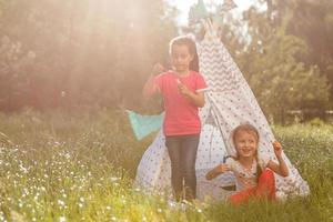 Zwei glückliche lachende kleine Mädchen im Campingzelt auf dem Löwenzahnfeld foto