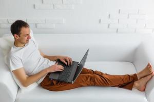 junger mann, der sich auf einem sofa mit einem laptop und einem handy entspannt foto