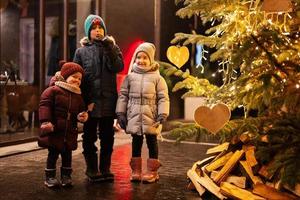 Drei Kinder amüsieren sich abends in der Nähe des beleuchteten Weihnachtsbaums im Freien. foto