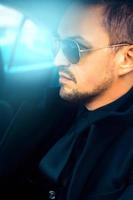 Wunderschöner erwachsener Mann im schwarzen Anzug und Sonnenbrille im Auto foto