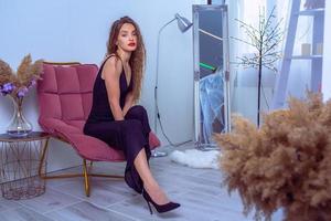 Glamour-Frau mit roten Lippen in schwarzen Overalls sitzt auf dem Stuhl und schaut in die Kamera foto