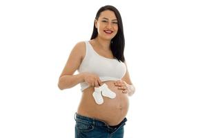 Studioporträt einer jungen schwangeren brünetten Frau mit kleinen Socken in ihren Händen, die vor der Kamera lächelt, isoliert auf weißem Hintergrund foto