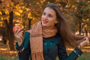 Schönes Mädchen mit langen Haaren und einem braunen Schal hält einen Apfel in der Hand und sieht weg, lächelt foto